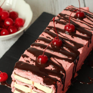 A white farmhouse table is laid with a stunning chocolate covered cherry ice cream sandwich cake and a bowl of maraschino cherries. The cake is on a black slate tray. You can see layers of ice cream sandwiches connected by cherry frosting. Chocolate sauce is drizzled on top and the cake is garnished with more cherries.