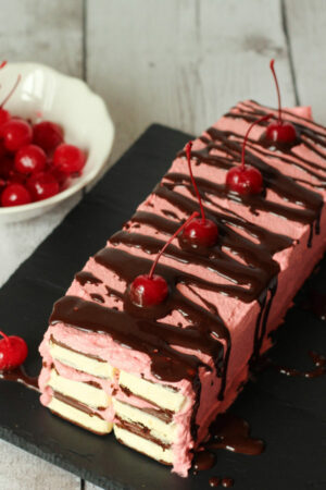 A white farmhouse table is laid with a stunning chocolate covered cherry ice cream sandwich cake and a bowl of maraschino cherries. The cake is on a black slate tray. You can see layers of ice cream sandwiches connected by cherry frosting. Chocolate sauce is drizzled on top and the cake is garnished with more cherries.