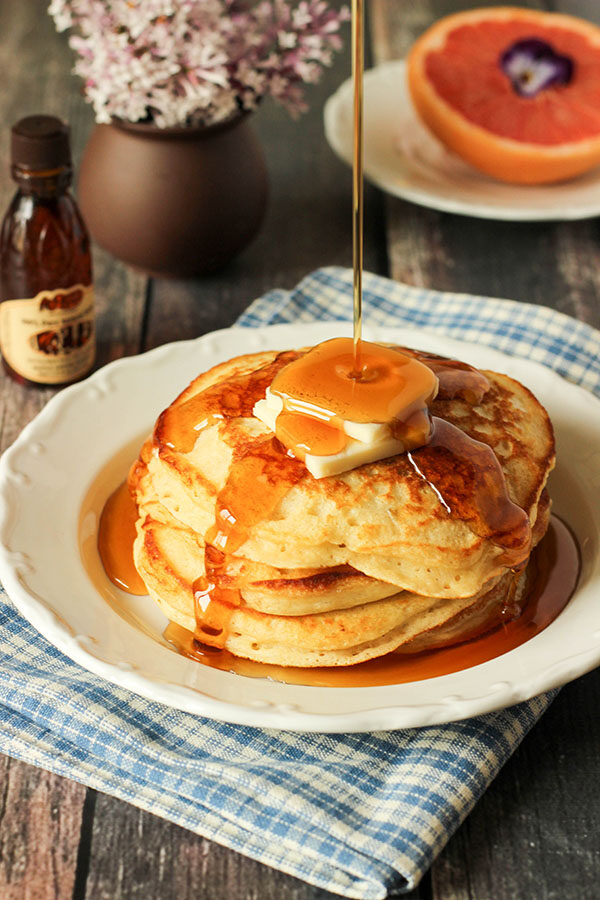 A lovely plate of copycat cracker barrel pancakes topped with syrup and butter. There is a vase of lilac in the background and half a grapefruit garnished with a pansy.