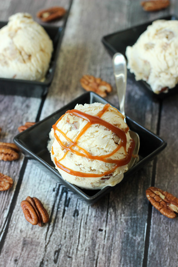 A big beautiful scoop of pralines and cream ice cream sits in a square black dish. It has salted caramel sauce drizzled over it and is surrounded by pecans.