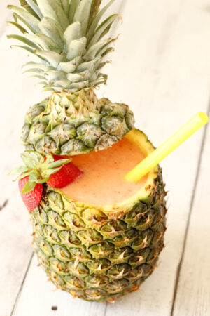 A pineapple is hollowed out and filled with cool, refreshing strawberry pina colada cocktail. There is a straw and a halved strawberry on the edge.