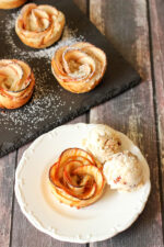 A beautiful apple pie rose sits on a white plate. There are scoops of ice cream on the side and a bit of caramel sauce drizzled over the top of the rose.