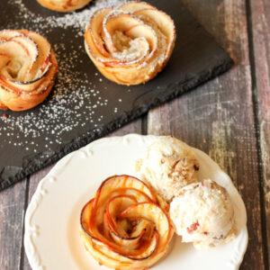 A beautiful apple pie rose sits on a white plate. There are scoops of ice cream on the side and a bit of caramel sauce drizzled over the top of the rose.