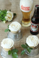 Table covered with burlap holds apple cupcakes frosted like white roses. There is a glass of apple beer nearby and each cupcake sits on a miniature overturned tin bucket.