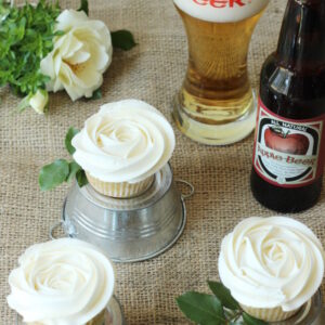 Table covered with burlap holds apple cupcakes frosted like white roses. There is a glass of apple beer nearby and each cupcake sits on a miniature overturned tin bucket.