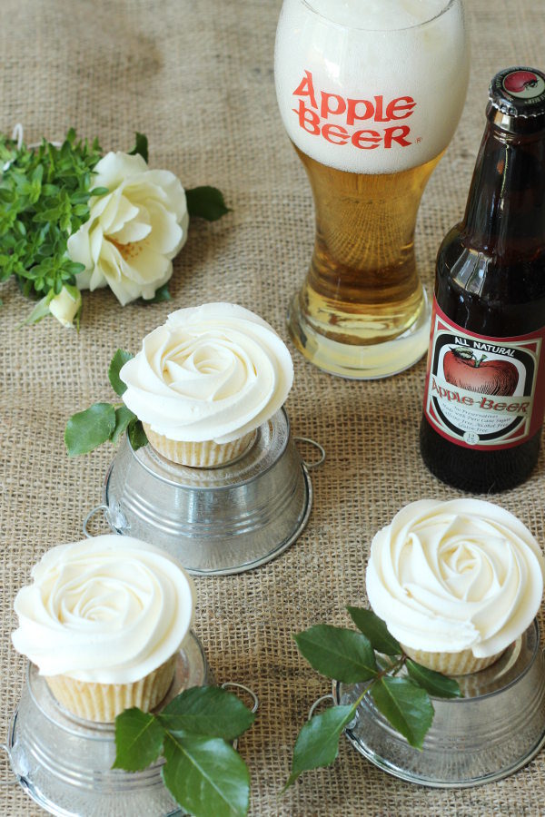 Table covered with burlap holds apple cupcakes frosted like white roses.  There is a glass of apple beer nearby and each cupcake sits on a miniature overturned tin bucket.