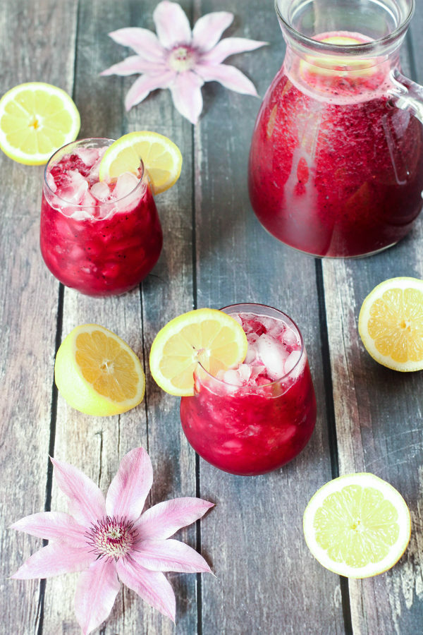 A gray wooden table is set with a pitcher of mixed berry lemonade that is a rich purple red from pureed berries. A few flowers and cut lemons sit nearby with stemless wine glasses of lemonade ready to drink.