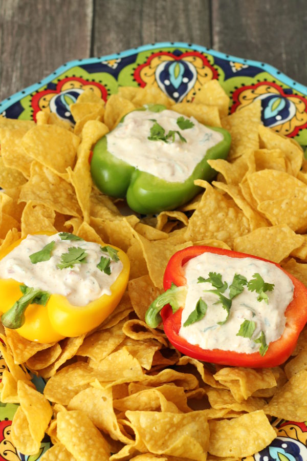 A big colorful platter is full of chips. Among the chips are three different colors of bell pepper and each pepper is cut in half and filled with a zesty, creamy Mexican dip. The dip is garnished with cilantro.