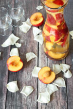A wooden table is covered with rose petals and nectarine halves. A carafe of white wine sangria full of sliced nectarines sits on the table by two empty glasses.