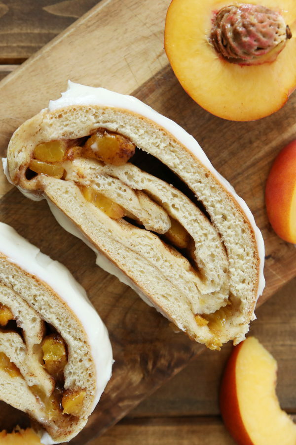 Two slices of cinnamon swirl peach bread are resting on a wooden cutting board. There is cream cheese glaze spread thickly on the top of the slices and some cut peaces around the bread.