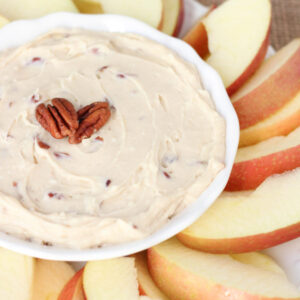 A white bowl of brown sugar apple dip sits in the middle of a platter of sliced apples. Pecans garnish the top.