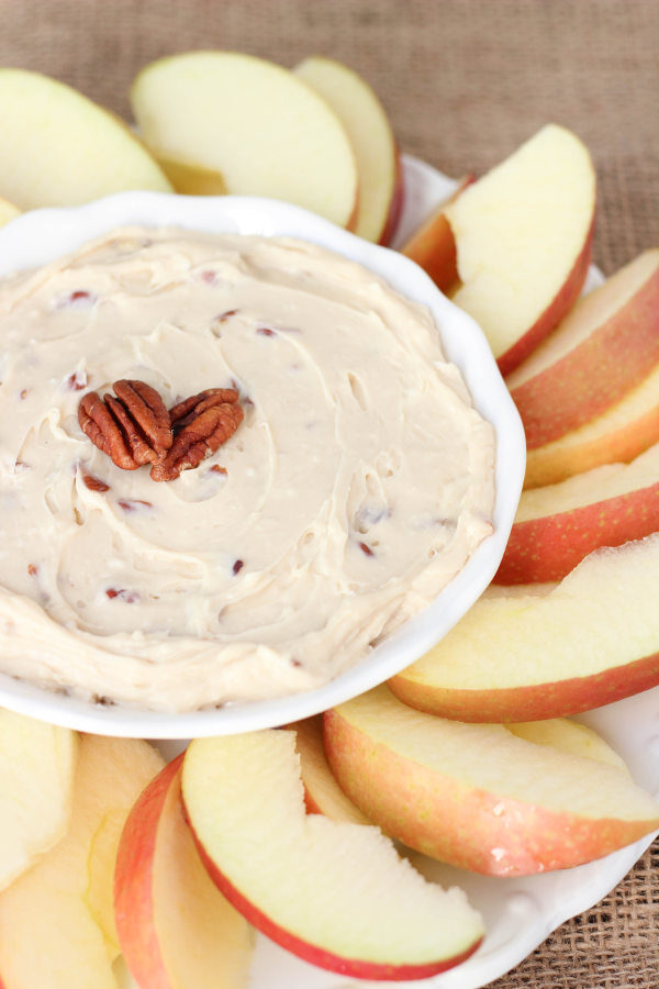 A white bowl of brown sugar apple dip sits in the middle of a platter of sliced apples. Pecans garnish the top.