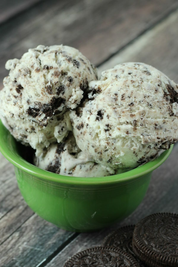 A green bowl is heaped high with big scoops of cookies and cream ice cream. You can see a few oreos on the gray table in front of the bowl.