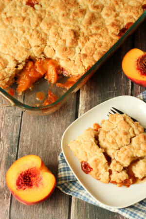 A big pan of easy peach cobbler has one slice cut out and plated on a white plate. Half a peach sits next to the dish.