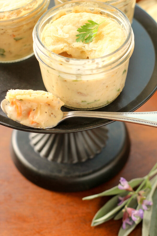 Three individual chicken pot pies baked in mason jars sit on a black stand.  You can see the puff pastry crust and bites of carrots and a creamy sauce.