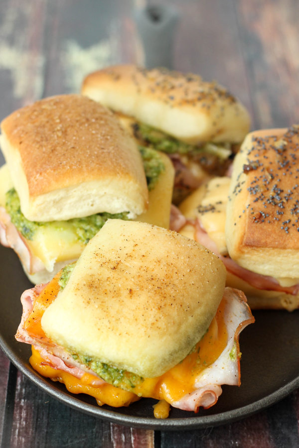 Four different hot ham and cheese sliders sit on a black plate. You can see ingredients like pesto and melted cheese and ham.