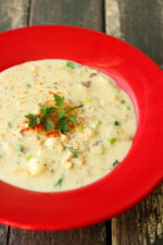 A bright red soup plate of clam chowder sits on a wooden table. The chowder is garnished with parsley and paprika.