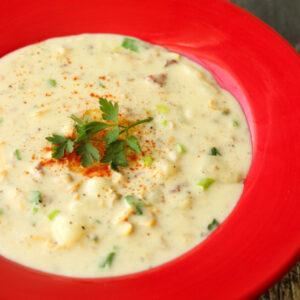 A bright red soup plate of clam chowder sits on a wooden table. The chowder is garnished with parsley and paprika.