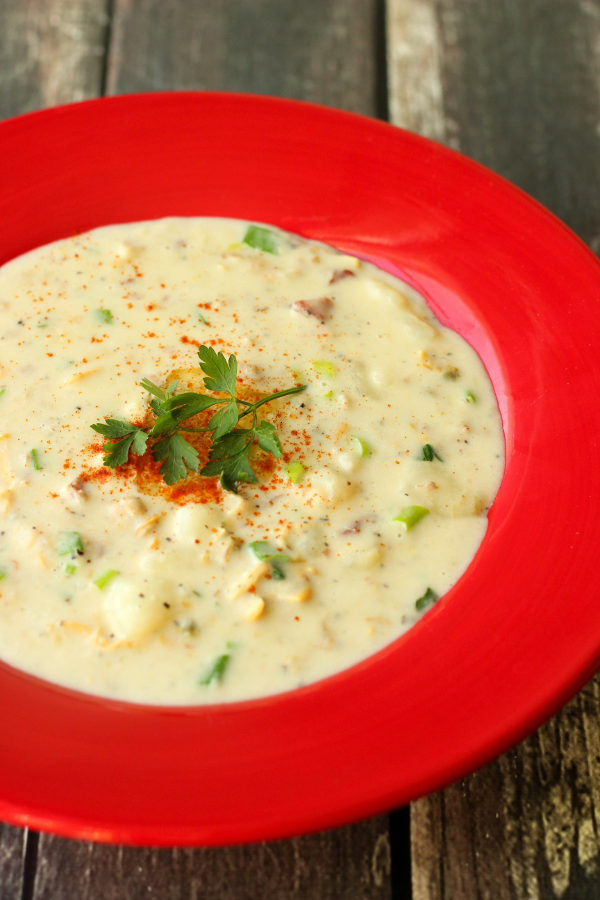 A bright red soup plate of clam chowder sits on a wooden table. The chowder is garnished with parsley and paprika.