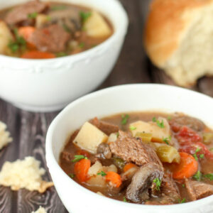 Two bowls of rich, filling beef stew sit on a table next to some torn bread. In the stew you see big chunks of beef, mushrooms, potatoes, carrots and some fresh parsley.