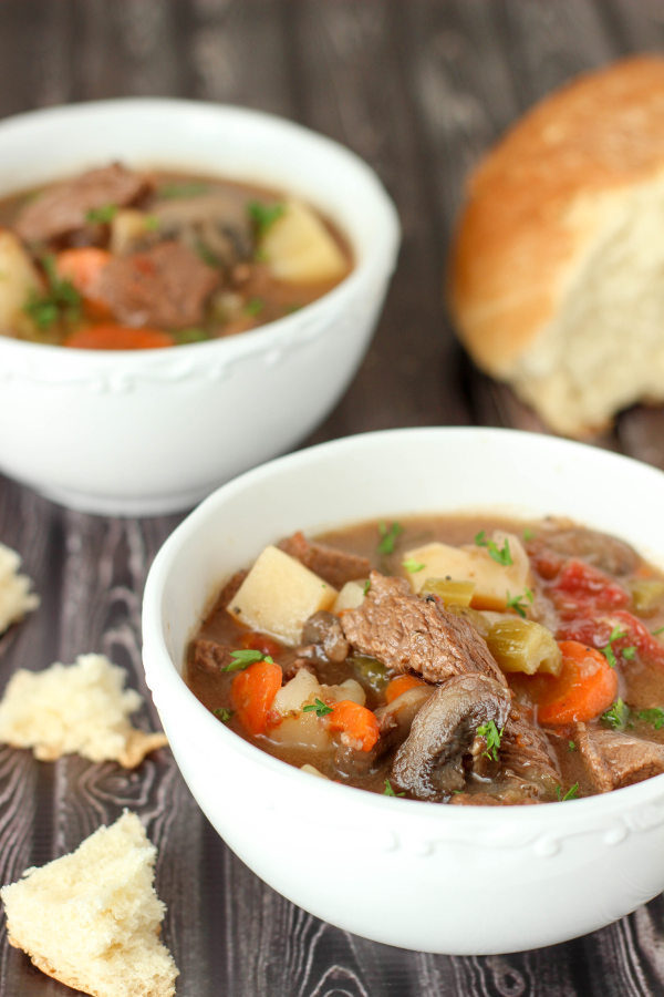 Two bowls of rich, filling beef stew sit on a table next to some torn bread. In the stew you see big chunks of beef, mushrooms, potatoes, carrots and some fresh parsley. 