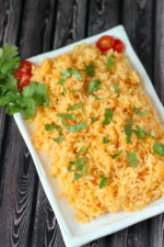 A white platter full of restaurant style Mexican rice sits on a dark wood table. There is a garnish of tomatoes and cilantro.