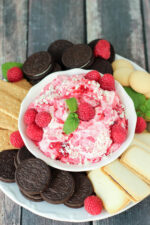 White chocolate raspberry cheesecake dip in a white bowl with fresh raspberries and mint on top. The bowl is surrounded by oreos, cookies, vanilla wafers, and graham crackers.