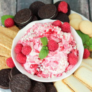 White chocolate raspberry cheesecake dip in a white bowl with fresh raspberries and mint on top. The bowl is surrounded by oreos, cookies, vanilla wafers, and graham crackers.