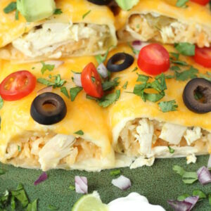 Two chicken and rice enchiladas sit on a green plate. They are covered with cheese and olives and tomatoes. Slices of avocado sit on the top.