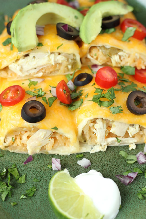 Two chicken and rice enchiladas sit on a green plate. They are covered with cheese and olives and tomatoes. Slices of avocado sit on the top.