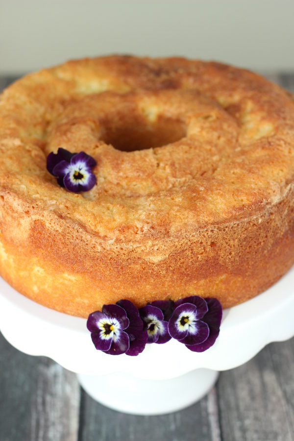 A beautiful pound cake sits on a white platter. There are purple pansies garnishing the cake.