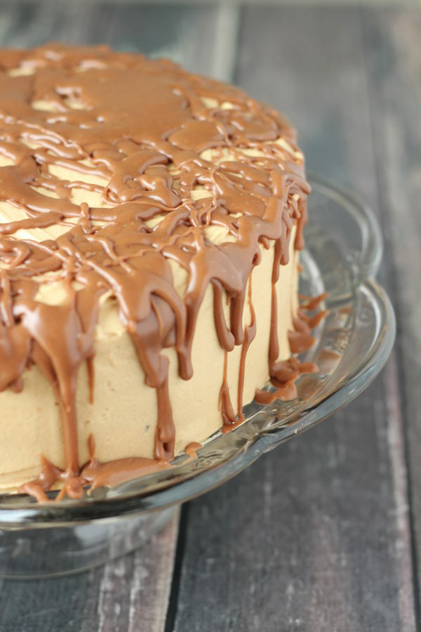 A chocolate cake covered with peanut butter frosting sits on a clear cake stand. The peanut butter frosting is covered with milk chocolate drizzle.