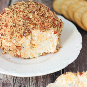 A gray farmhouse table is set with a big classic cheese ball on a white plate. The cheese ball is rolled in crushed smoked almonds and there are ritz crackers handy.