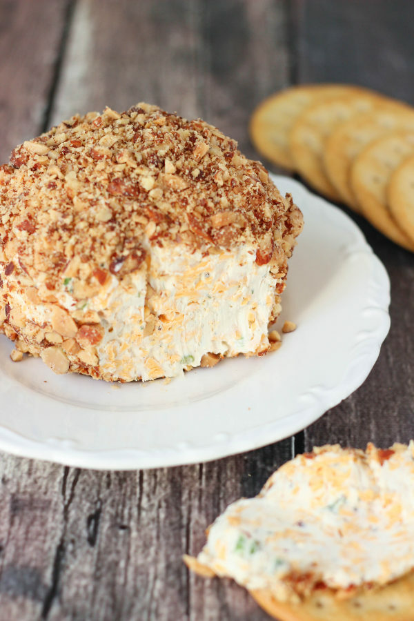 A gray farmhouse table is set with a big classic cheese ball on a white plate. The cheese ball is rolled in crushed smoked almonds and there are ritz crackers handy. 