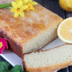This drizzled lemon loaf is on a white plate. Fairy daffadills are on top and lemons and lemon balm garnish the loaf.