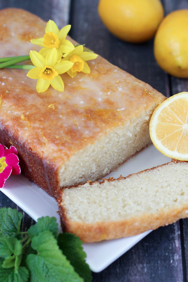 This drizzled lemon loaf is on a white plate. Fairy daffadills are on top and lemons and lemon balm garnish the loaf.