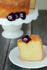 A large round pound cake sits on a white cake stand. A slice is cut out and sitting in front of the cake on a white plate. Pansies garnish both.