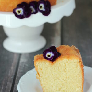 A large round pound cake sits on a white cake stand. A slice is cut out and sitting in front of the cake on a white plate. Pansies garnish both.