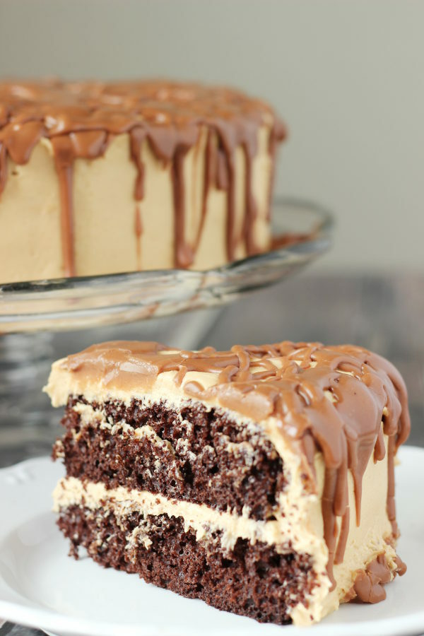A big wedge of chocolate cake is covered in peanut butter frosting and then drizzled with milk chocolate drizzle. The rest of the cake is on a clear glass cake plate in the background.