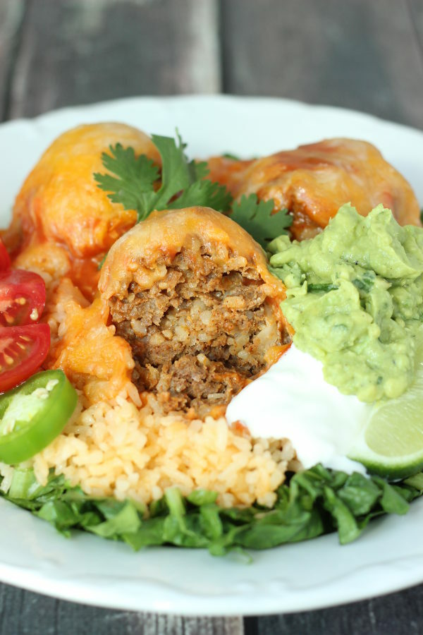 A platter of taco balls with all the fixings. There are three taco meatballs covered in cheese. They are plated with lettuce, rice, sour cream, guacamole, and tomatoes.