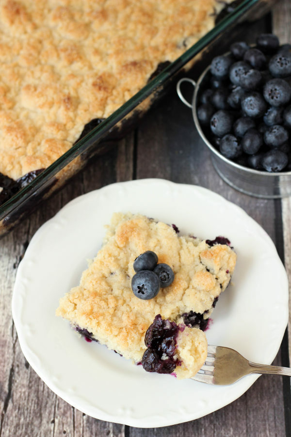 A white plate holds a big square of blueberry cobbler topped with a few fresh blueberries. In the background you see a cooked blueberry cobbler and a tin pail overflowing with blueberries!