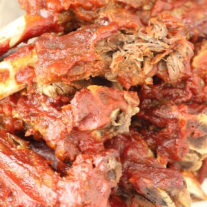 A big pile of bbq beef ribs is stacked up on a plate. The ribs are covered in a spicy red sauce.