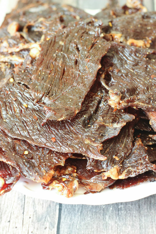 A big pile of strips of beef jerky are stacked on a white dinner plate. You can see black and red pepper on the jerky.