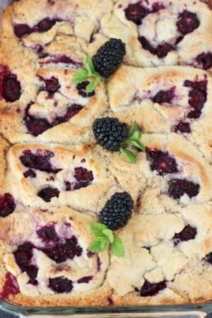 A big pan of blackberry cobbler sits waiting to be eaten. There are fresh blackberries and mint leaves on top.