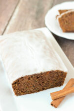 A fresh, tender loaf of carrot and zucchini spice bread sits on a white plate. The loaf is covered in apple cider glaze and sitting next to some cinnamon sticks. A plate of slices is ready to eat in the background.