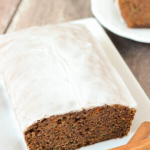 A fresh, tender loaf of carrot and zucchini spice bread sits on a white plate. The loaf is covered in apple cider glaze and sitting next to some cinnamon sticks. A plate of slices is ready to eat in the background.