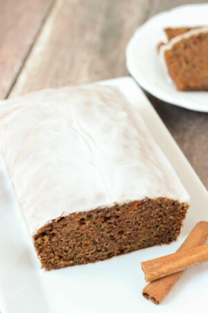 A fresh, tender loaf of carrot and zucchini spice bread sits on a white plate. The loaf is covered in apple cider glaze and sitting next to some cinnamon sticks. A plate of slices is ready to eat in the background.