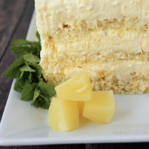 A simple pineapple cake sits on a white platter. There are three layers of pound cake and three layers of pineapple cream full of crushed pineapple. The plate is garnished with a sprig of mint and three chunks of pineapple.