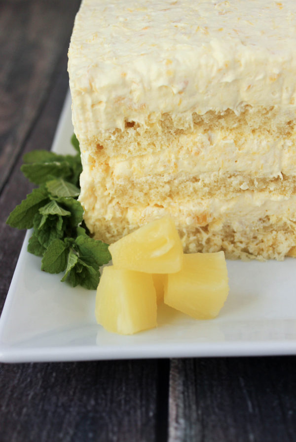 A simple pineapple cake sits on a white platter. There are three layers of pound cake and three layers of pineapple cream full of crushed pineapple. The plate is garnished with a sprig of mint and three chunks of pineapple. 