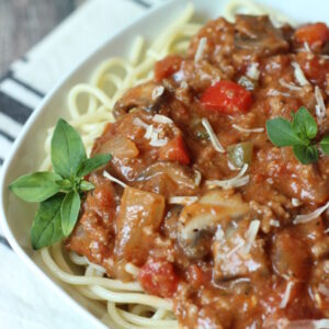 A big square bowl is full of spaghetti noodles and topped with a meaty, chunky spaghetti sauce. There are some basil leaves as garnish and a cloth napkin on the table ready to sop up some tomato sauce.